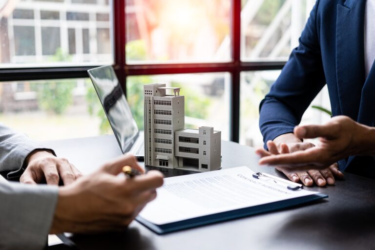 A close-up shot shows two people at a desk, facing each other, with a miniature white building model between them. One person, on the left, is wearing a light-colored suit jacket and is holding a pen, poised to write on a document in a dark blue folder. Their left hand is resting on the document. The other person, on the right, is wearing a dark blue suit jacket, and their hands are visible, gesturing as if in conversation.